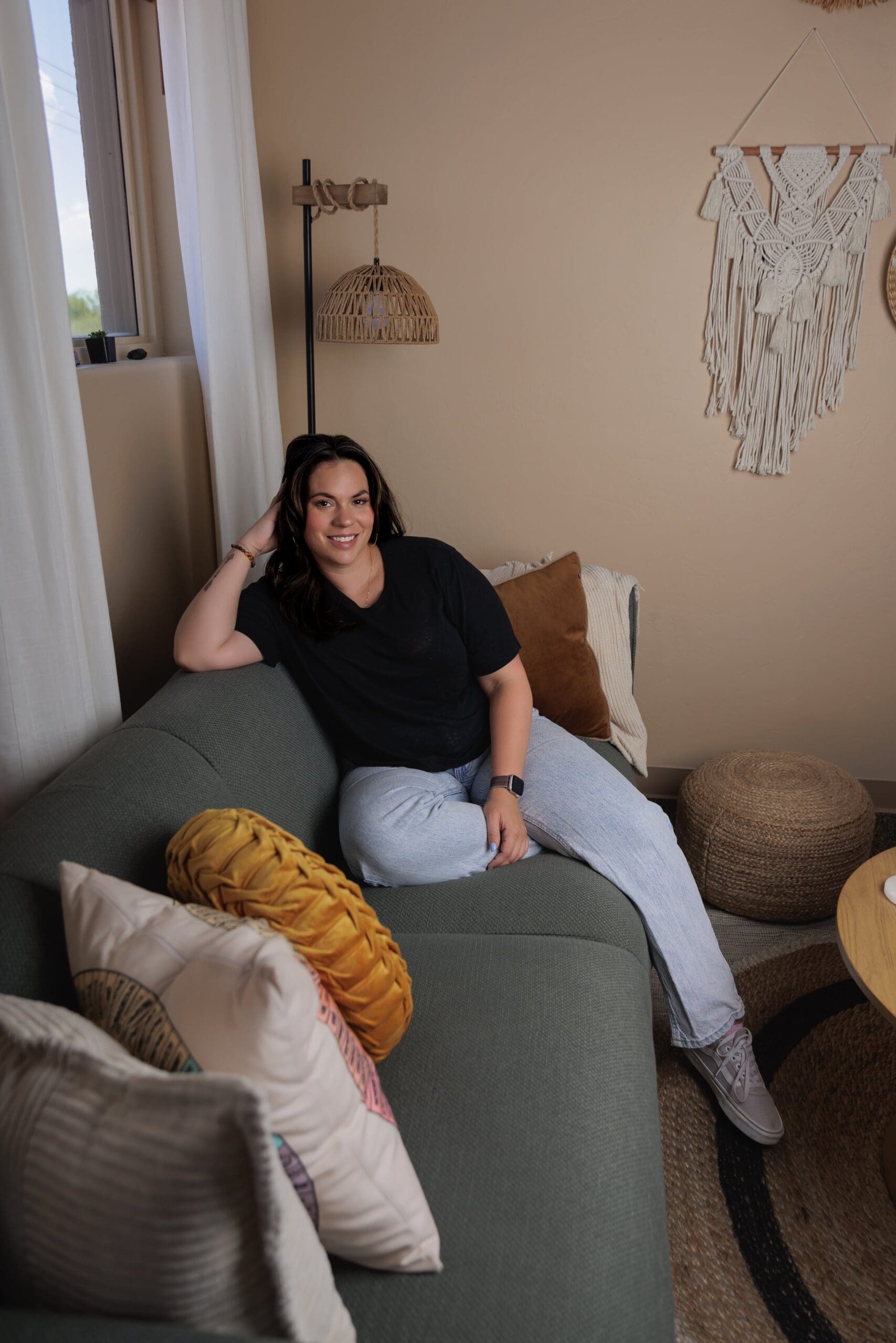 Stephanie McInelly sitting on a gray couch, dark shirt and light denim jeans, arm propping her head up, smiling